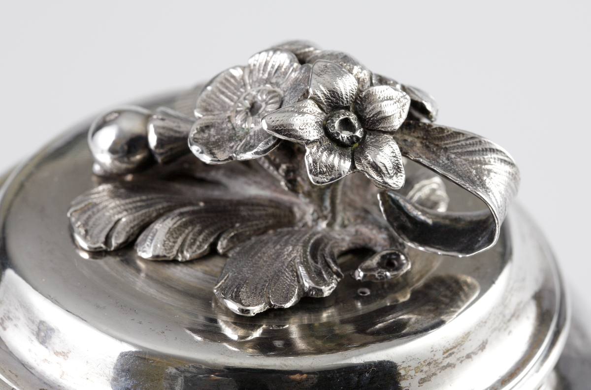 Kaffekanna i silver. 
Cylindrisk modell på fyra tassfötter. Pärlstavsdekor upptill och nedtill, innanför denna en ciselerad palmettbård. Välvt lock med graverad bård och knopp i form av blombukett. Kort, rakt utstående holk för skaftet. Vid holkens mynning dekor av pärlbård, vid dess inre kortsida omgiven av en pålödd och ciselerad blomma. 
Försedd med svarvat, svärtat trähandtag, fäst med ett bladformat böjt silverstift (saknas).
Stämplar på undersidan av kannan.

Inskrivet i huvudbok 1952.