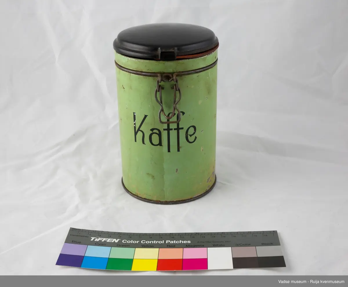 Sylinderformet kaffeboks i metall med hengslet plastlokk. Enkel låsemekanisme i metall i front. Lokket er i brun plast og utstyrt med en gummipakning. Boksen er helfarget grønn utvendig, med påskriften "Kaffe"i svart i front.