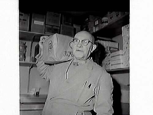 John Johansson står vid en trave ägg i Konsum Frillesås. I bakgrunden står rullar med vad som kan vara prover på olika linoleummattor.
Bild 2 visar John med en i papp inslagen sockerpaket på axeln.