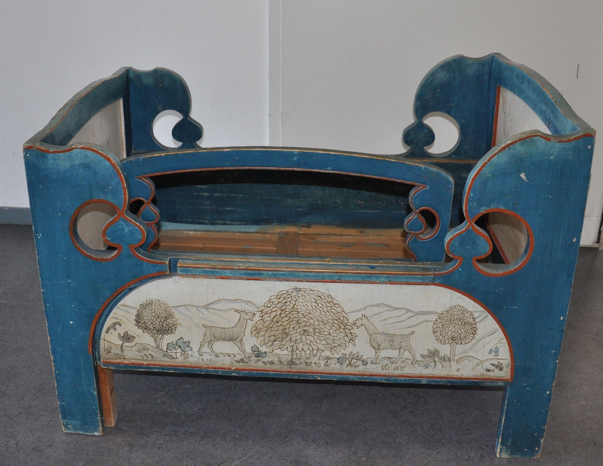 Uttrekkbar barneseng av tre, i to deler. Sengen er malt blå med dekorative røde linjer ved kanter. Hode- og endegavel har kurvet form øverst, og fylling på utsiden. Det er malt dekor på utsiden av endegavel og sideplater, og på innsiden av hodegavel. Motiv på maling av to fugler og sol på hodegavel, to trær og Sør-Fron kirke på endegavel og kuer og trær på sidegavler. Spesielt på hodegavel er malingen avlitt. Det følger med en løs sengehest med sveifet dekor.