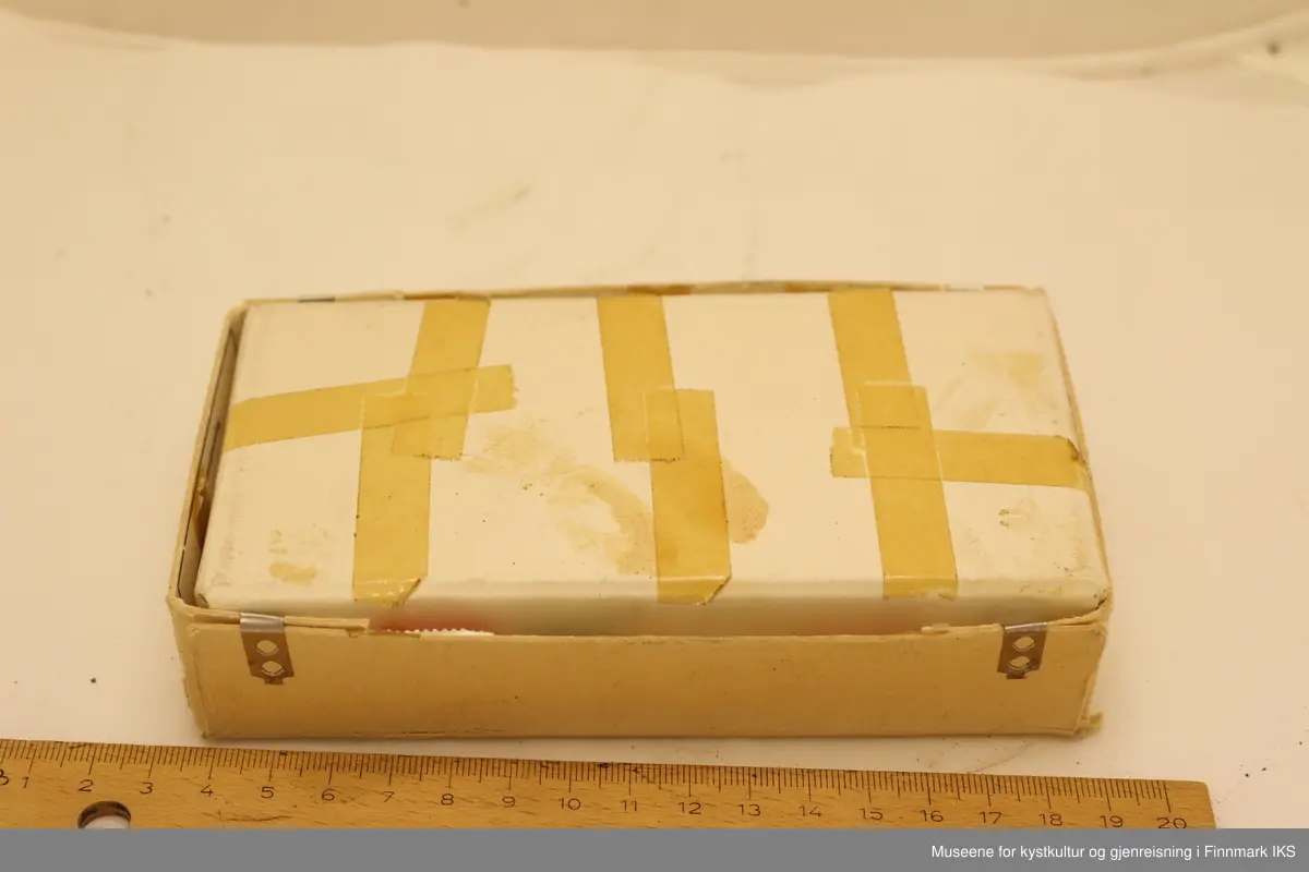 Briller i post-pakke som fungerer som brilleetui. Brilleetuiet består av en avlang firkantet boks med bunn og topplokk, og inneholder regning, poststempel og frimerker samt adresse til mottager og returadresse på undersiden av topplokket. Brillene er laget i gull- og brunfarvet plast og gullfarvet metall, mens boksen er laget i papp.