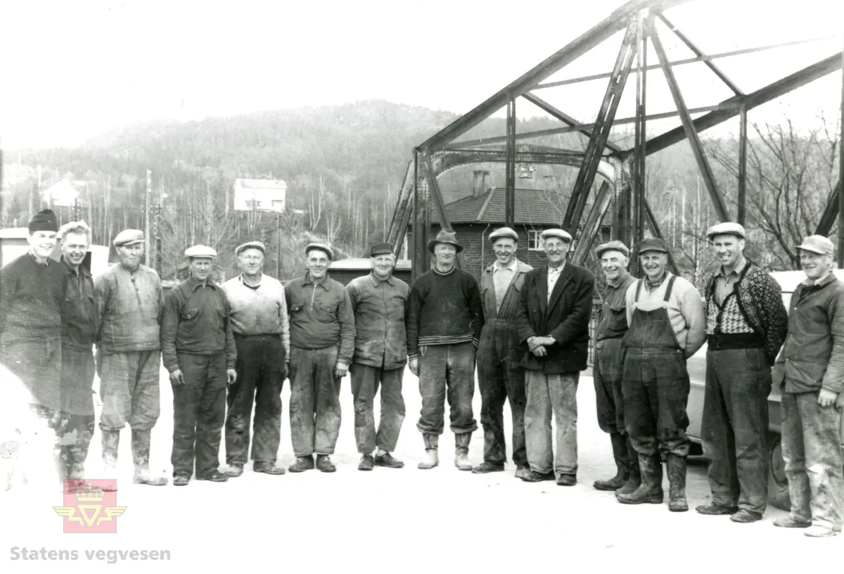 Arbeidere oppstilt ved Hokksund bru. 
Fra venstre:  Kjell Bollerud, Rolf Øderud, Ingvald Knivebakken, Hans Haare, Oskar Hvam, Karsten Berg, Johan Johannesen, Ole Engerud, Lyder Marstein, Oskar Ombye (formann), Ottar Hvidsten, Trygve Tiedeman, Karsten Bogen og Asbjørn Andersen. Ikke tilstede: Sigurd Leversby, Henry Thon. 
Formannen på bildet heter Oskar  Ombye. 

Informant: Nina Kristiansen (barnebarn)
Informant: Ulf Kristian Nilsen (barnebarn)