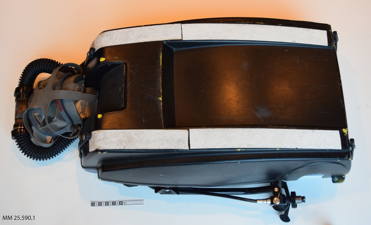 Dykapparat med andningsmask. Apparaten är fastsatt inom ett plasthölje vars form påminner om en resväska. Plasthäöjet öppnas opch stängs med hjälp av gummistroppar med hål som träs över skruvar/nitar. Inuti höljet finns två flaskor, en blå och en grön (doserflaska och blandgasflaska). Fäst i den blå flaskan löper två svarta gummislangar som går till andningsmasken. Masken är av grått gummi och täcker bärarens hela ansikte. Masken träs vöer ansiktet och runt bakhuvudet på bäraren sitter fyra gummidelar som förenas i en platta av gummi.