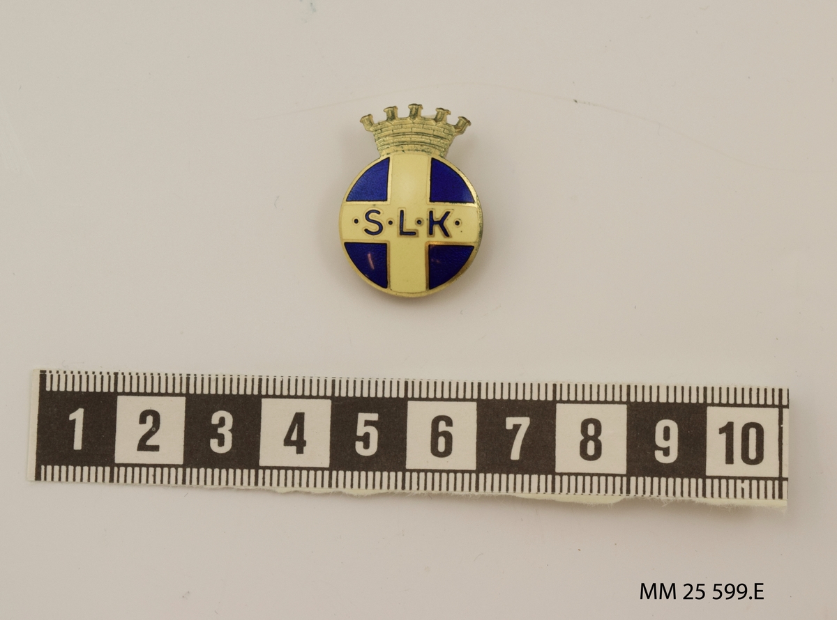 Lottamärke av metall. Cirkelrunt märke med krona i guld över. Emaljerat motiv av svenska flaggan, blå bakgrund med gult kors. Bokstäverna "SLK" i blått i korsets mitt. Baktill en nål för fästning på kläder. Märkning i relief: "Sporrong & Co, Stockholm".