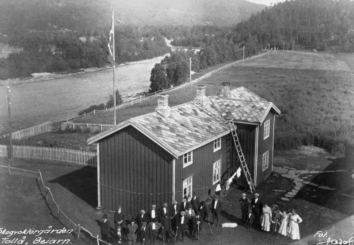 Skogvoktergården Tollå i Beiarn. Fotografiet er tatt fra et høyt punkt, muligens et hustak, mot skogvokterboligen.  Dette er en toetasjes, rødmalt bygning med vertikalstilt, antakelig rødmalt panel.  Vindusomramminger hjørnebord og vindskier er kvitmalte, og taket har skifertekke.  Bygningen har en bakstueløsning i to etasjer som har saltak vinkelrett på hovedfløyens møneretning.  Til venstre foran huset står 17 velkledde herrer, flere av dem med spaserstokker, antakelig deltaktere i et skogfaglig møte. Til høyre for denne gruppa ses to herrer, fire kvinner og to barn, som antakelig har betjent møtet.  Bakenfor bygningen er det en liten hage med flaggstang, inngjerdet av et kvitmalt stakitt.  Bakenfor er et forholdsvis flatt, grasbevokst åkerareal og ei stilleflytende elv, samt en del skog. 