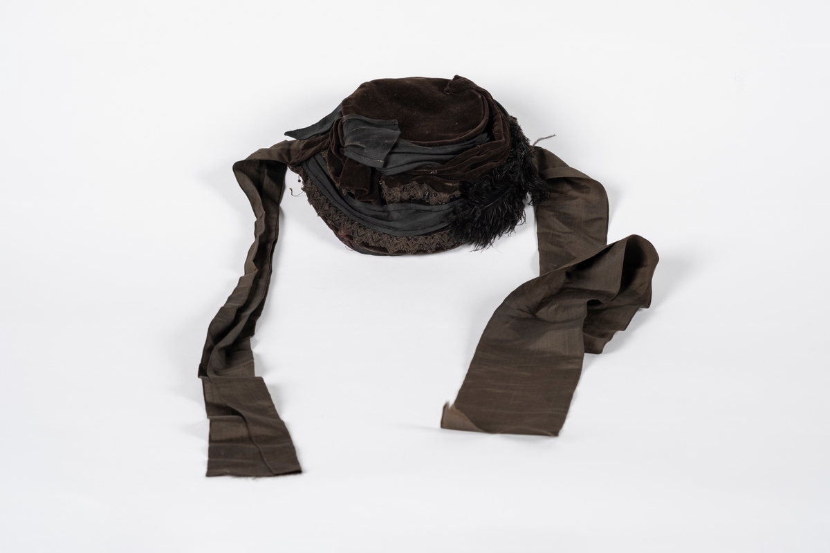 Rund hatt av plast trukket med fløyel. Hatten er pyntet med blondebånd, perler og fjær. Pullen er drapert med fløyel og ripsvevet bomull som ender i en sløyfe. Hatten har hakebånd. Svettebåndet er plastbelagt.