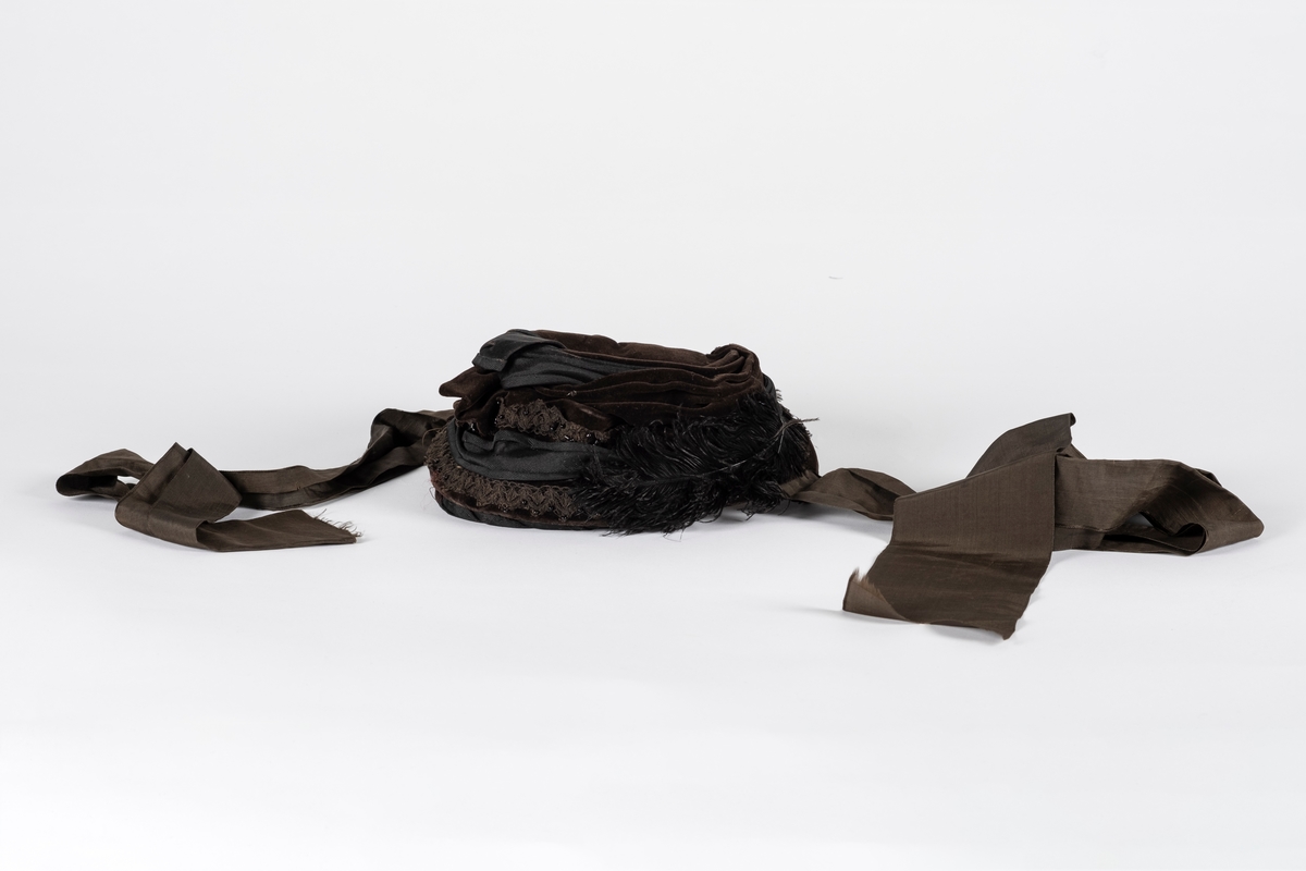 Rund hatt av plast trukket med fløyel. Hatten er pyntet med blondebånd, perler og fjær. Pullen er drapert med fløyel og ripsvevet bomull som ender i en sløyfe. Hatten har hakebånd. Svettebåndet er plastbelagt.