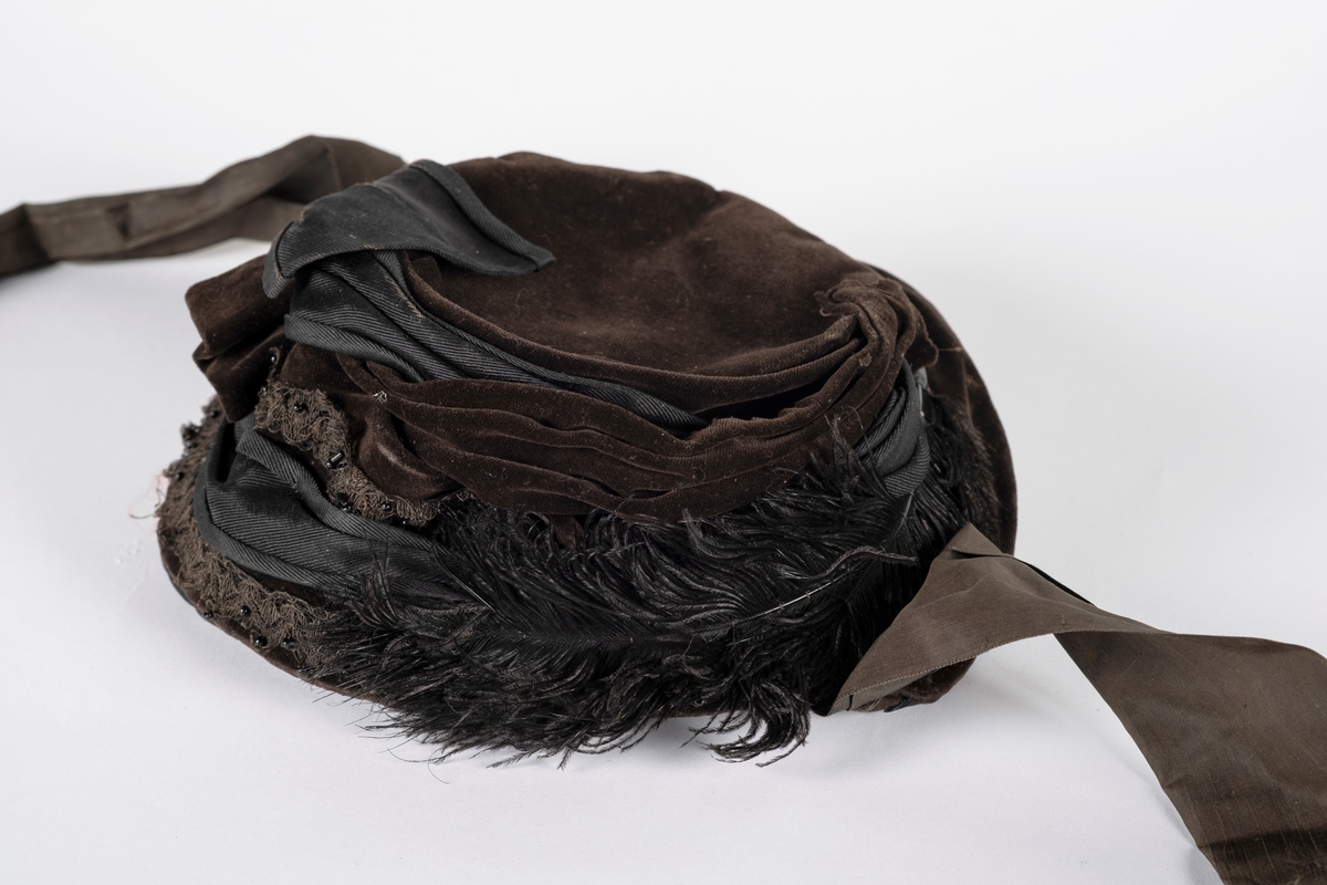 Rund hatt av plast trukket med fløyel. Hatten er pyntet med blondebånd, perler og fjær. Pullen er drapert med fløyel og ripsvevet bomull som ender i en sløyfe. Hatten har hakebånd. Svettebåndet er plastbelagt.