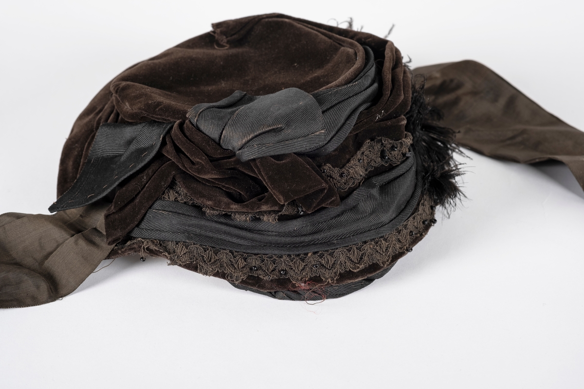 Rund hatt av plast trukket med fløyel. Hatten er pyntet med blondebånd, perler og fjær. Pullen er drapert med fløyel og ripsvevet bomull som ender i en sløyfe. Hatten har hakebånd. Svettebåndet er plastbelagt.