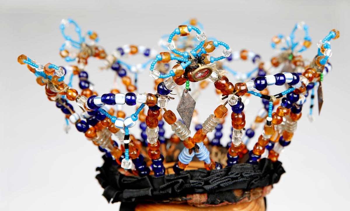 Kronen er laget av ståltråd påtredd glassperler i ulike fasonger og farger. Den består av tolv buer som står opp som går om hverandre. På toppen av annenhver bue er det laget tre mindre buer av mindre perler med innslag av større. Under et par av disse er det festet blanke knapper. Det sitter igjen heng av metall under fire av disse buene. På de andre buene er det festet heng av mindre perler med større perler på midten hvorfra det er heng av mindre perler med en større nederst.. I en av disse hengene skiller perlene seg ut fra de andre.. Krona har valk surret med, og påsydd, bånd og svart stoff. Utenpå valken er det feste et ripsbånd som er foldet til pynt. Under valken er det feste tråd i kryss. Krona har knyting i svarte ripsbånd som er sydd fast dobbelt.
