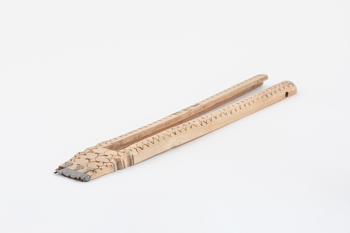 Del av vevspenne av tre, ytterdelen. Vevspenna har utskåret bølgeformet dekor, samt utskjæring i den ene enden. Det er et beslag av metall i den ene enden med pigger.