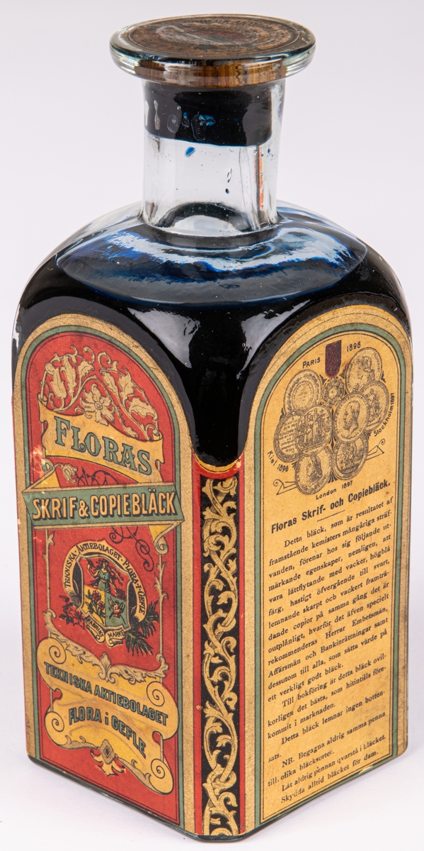 Bläckflaska med innehåll. Av glas. Oöppnad. Etiketter på samtliga sidor, varav framsidans i rött och med texten "FLORAS SKRIF & COPIEBLECK - TEKNISKA AKTIEBOLAGET FLORA I GEFLE", m.m.
Kvadratisk genomskärning med fasade hörn. Rundas upp till en smal rund hals med horisontell krage överst. Förseglad med pappetikett över korken. Etiketter på samtliga sidor, varav framsidans i rött med detaljer i flera färger. Sidoetiketterna har längre beskrivningar av bläcket på svenska resp. franska. På baksidan räknas prisbelöningar upp.