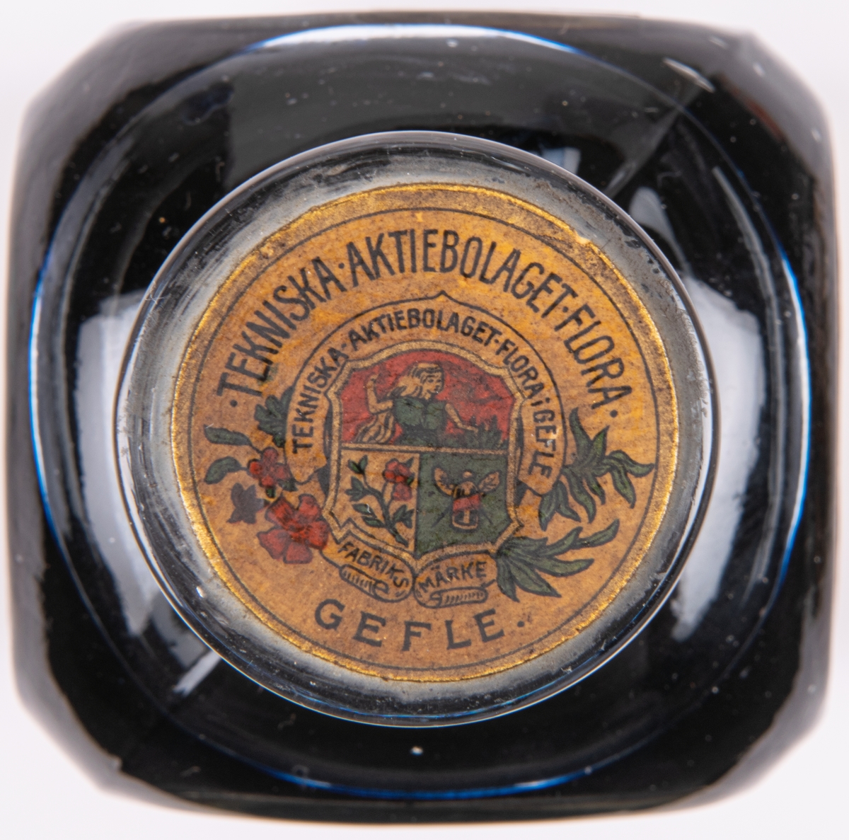 Bläckflaska med innehåll. Av glas. Oöppnad. Etiketter på samtliga sidor, varav framsidans i rött och med texten "FLORAS SKRIF & COPIEBLECK - TEKNISKA AKTIEBOLAGET FLORA I GEFLE", m.m.
Kvadratisk genomskärning med fasade hörn. Rundas upp till en smal rund hals med horisontell krage överst. Förseglad med pappetikett över korken. Etiketter på samtliga sidor, varav framsidans i rött med detaljer i flera färger. Sidoetiketterna har längre beskrivningar av bläcket på svenska resp. franska. På baksidan räknas prisbelöningar upp.