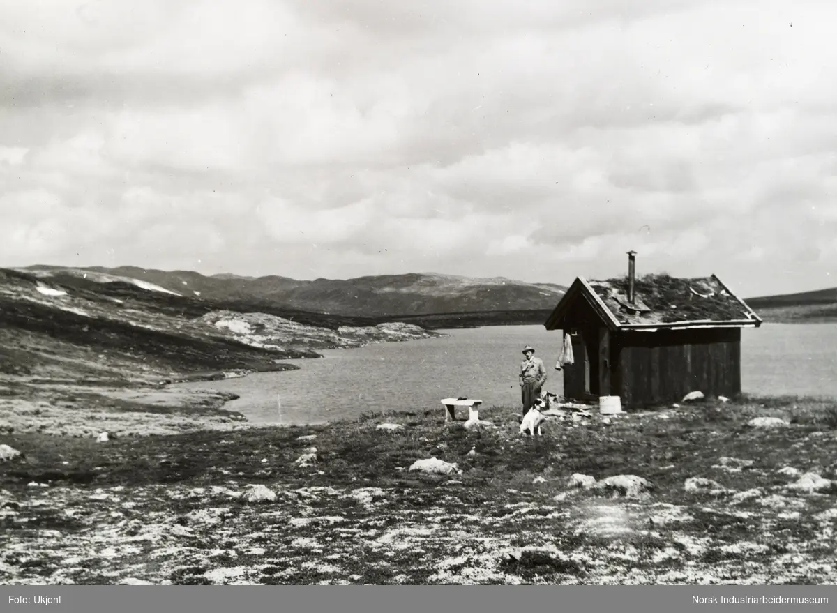 Mann og hund ved liten hytte. I bakgrunn sees innsjø og fjell