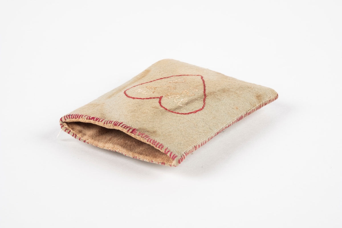 En liten firkantet lomme laget av papp kledd med tekstil. Inne i lommen er det en papirlapp. Det er brodert ruter med årstall under på den ene siden og et hjerte med bokstaver i på den andre.