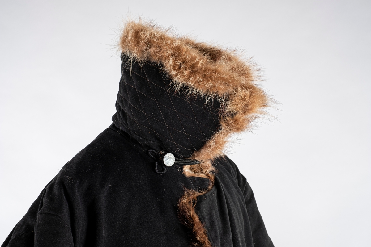 Frakk av bomull med pelsforing. Den har en bred krage. Kragen og mansjettene er kledd med pels. Frakken kneppes i halsen med knapp og hempe på hver side. Ved nedre kant er det brodert 3 røde kryss.
