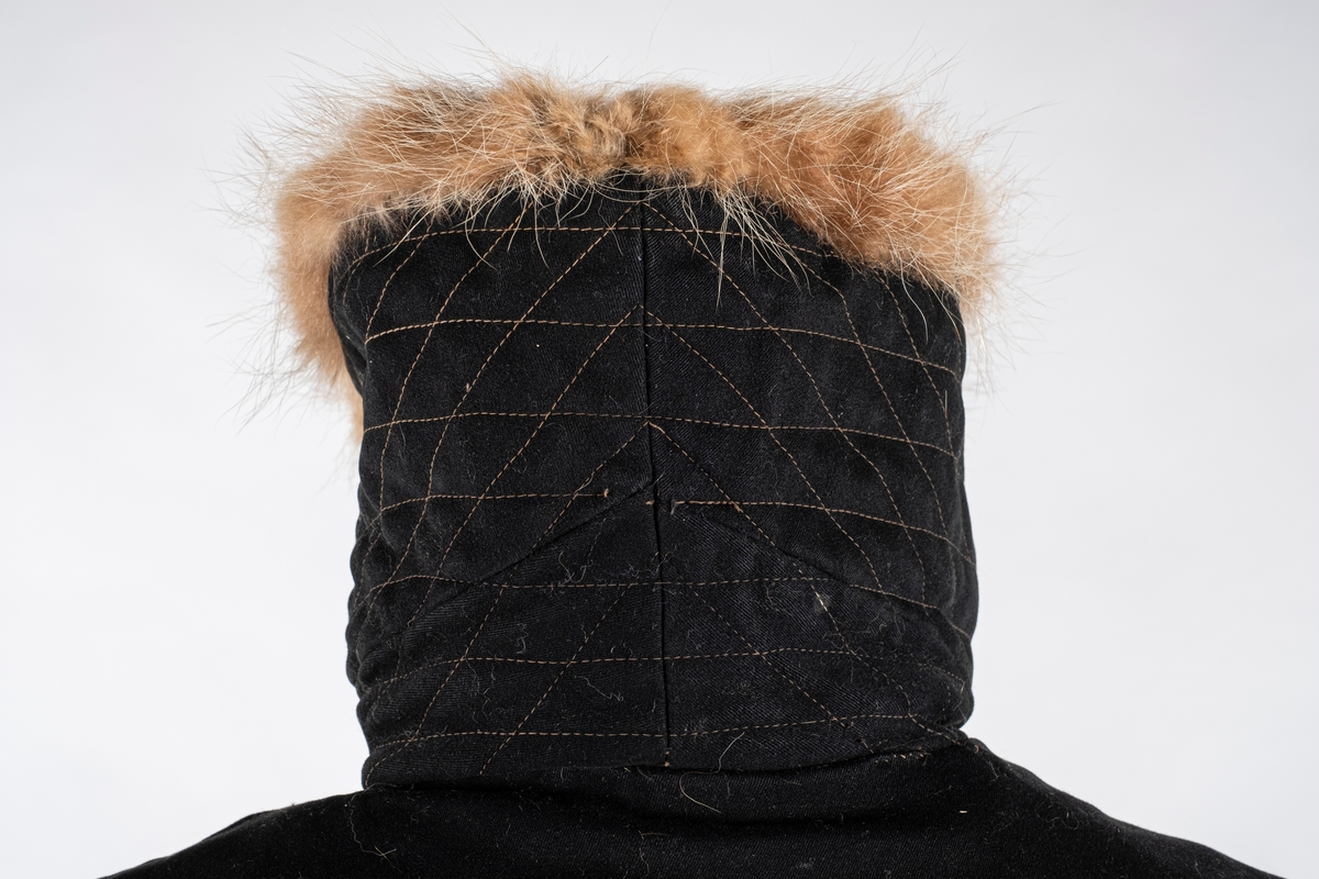 Frakk av bomull med pelsforing. Den har en bred krage. Kragen og mansjettene er kledd med pels. Frakken kneppes i halsen med knapp og hempe på hver side. Ved nedre kant er det brodert 3 røde kryss.