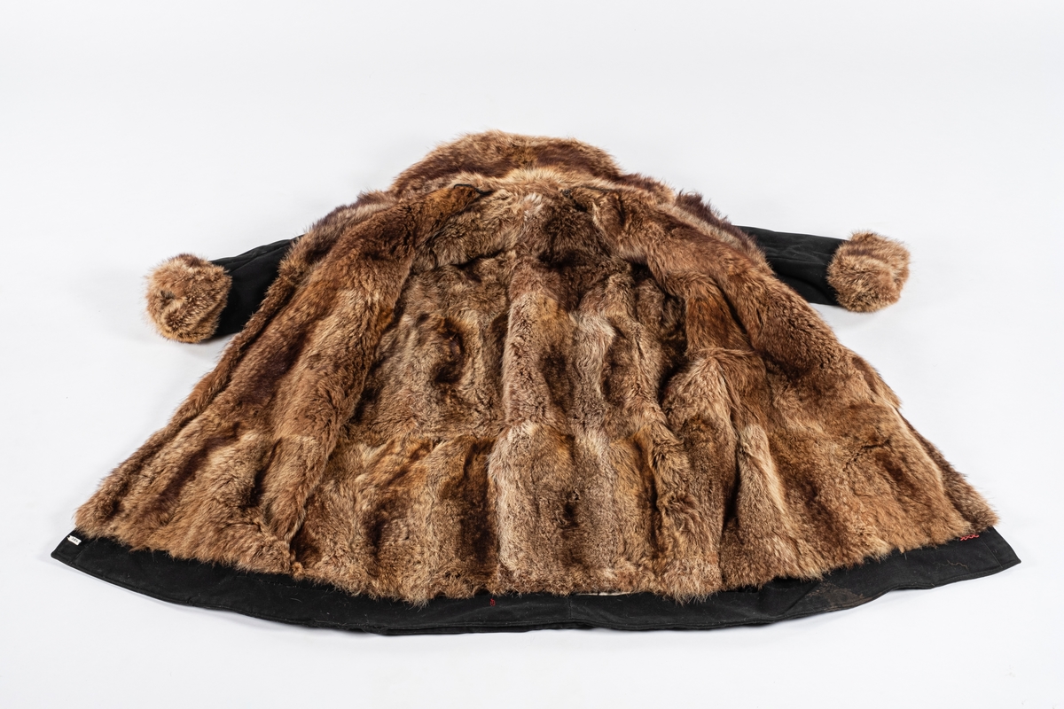 Frakk av bomull med pelsforing. Den har en bred krage. Kragen og mansjettene er kledd med pels. Frakken kneppes i halsen med knapp og hempe på hver side. Ved nedre kant er det brodert 3 røde kryss.