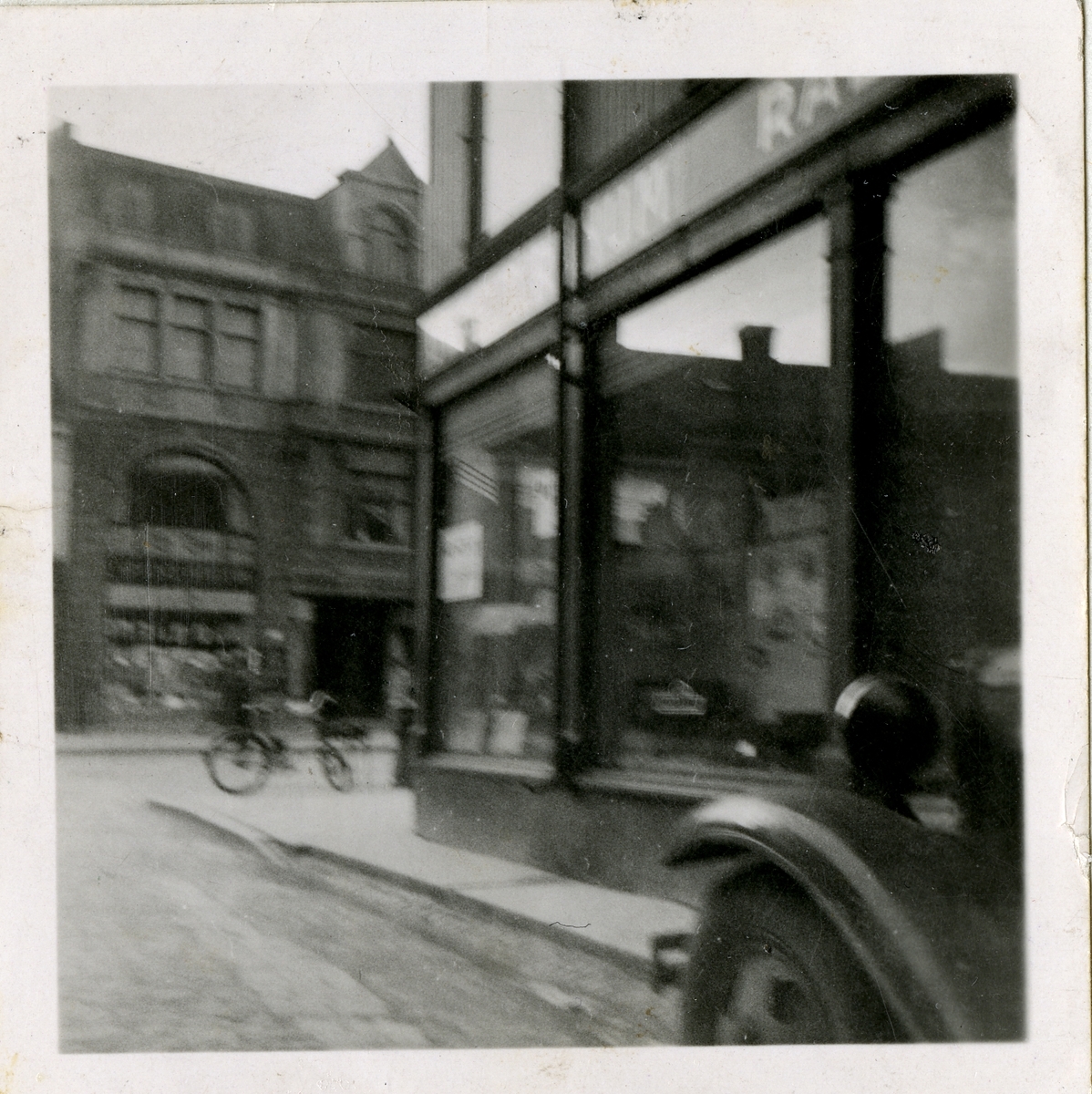 Fra familiealbum. Fotografi tatt fra gaten med Olaf T. Ranum A/S' butikkfasade på høyre side og på motsatt side nummer 21 (murbygningen) Kioskenes ekspedition i Olav Tryggvasons gate, Trondheim.