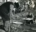 Trumslagarbacken, Västerås.
Arbetsbild från Vallbygrävningen 1958.
Äldre man i väst, glasögon och hatt mäter något med måttband.
Yngre man i rutig skjorta sitter vid provisoriskt ritbord och skriver troligen in måtten på karta.