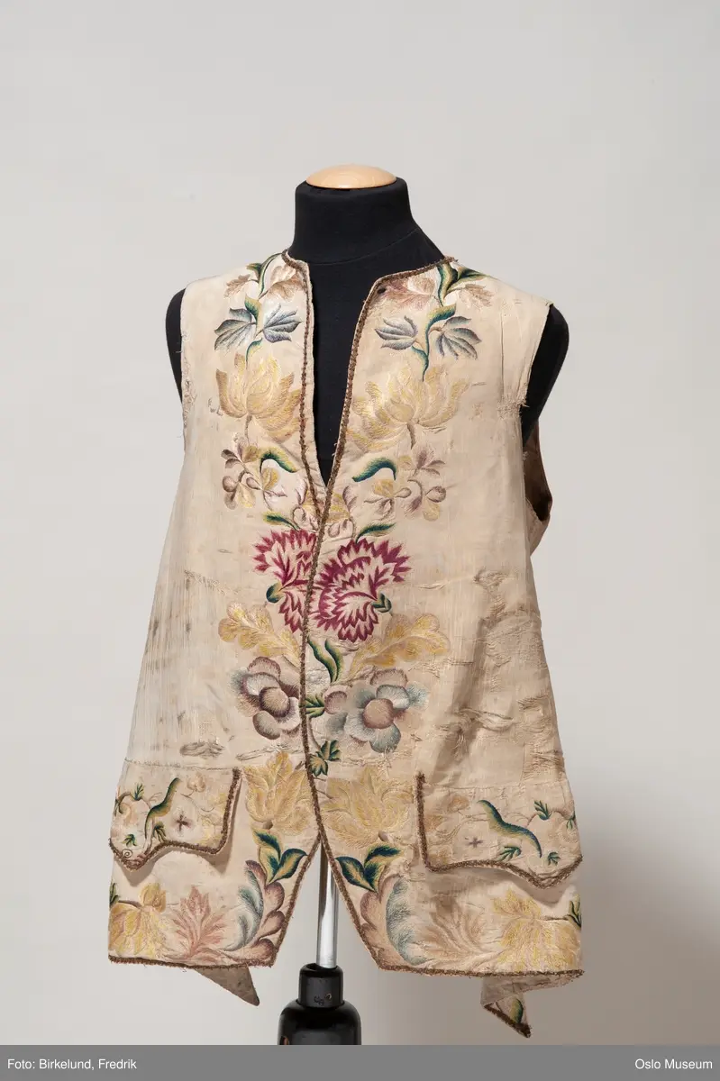 Lang 1700-talls vest, kortere bak. Kremgul silke, brodert (gult, grønt, rødt, brunt). Kantet med falmet gull bånd, også på lommenes slag. Fire knapper. Bomull i bakstykke , snøring i bak med hvitt bomullsbånd.