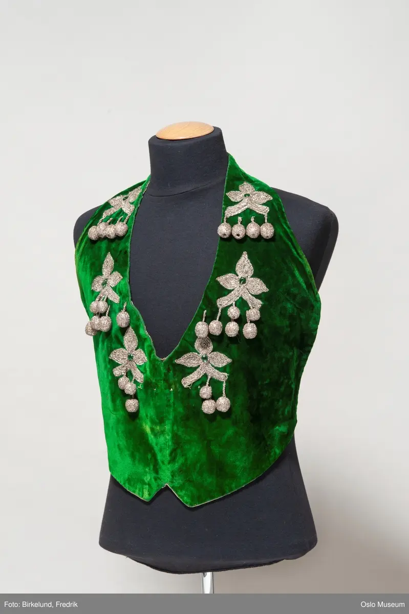 A: vest, grønn bunn med broderte blomster i metallfarget tråd. Pyntet med rød plaststeiner. 
B: bukse, grønn bunn med blomster brodert i metallfarget tråd langs kanten. Rosa dekor. 
C: skjorte, hvit lin. 
D: løse deler
E: vest