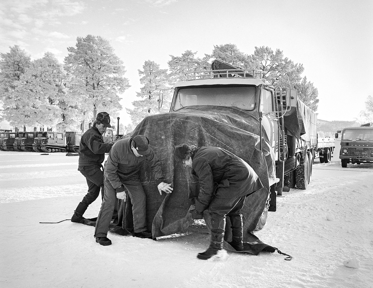 Sveg 1982.

Efter att snabbmotorvärmaren monterats täcks motorhuven med presenning. Mellan framhjulen monteras också en tillpassad bit presenning.
Kanterna mot marken täcks med snö -- allt för att bibehålla värmen från motorn så länge som möjligt.
I mitten ser vi teknikläraren 1. ding Rolf Nordström.

Det fungerar! Har praktisk erfarenhet, bland annat från Sveg januari 1966 då termometern visade -42 grader./P-O Rosén
