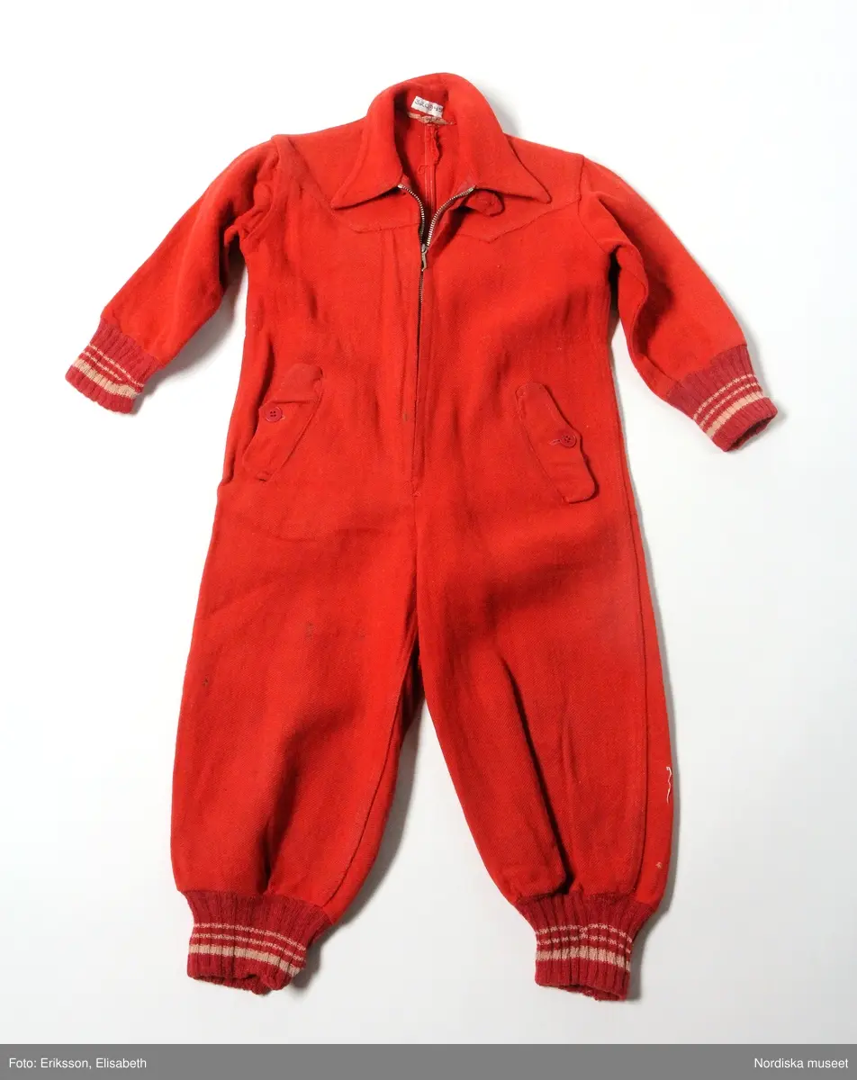Katalogkort:
"Overall av rött kypertvävt ruggat ylletyg. 
Två framstycken, två bakstycken, fram besparing med snibbig nederkant. Knäppning mitt fram med långt blixtlås av metall (Wico), överst slejf med knapphål samt röd knapp, krage med spetsiga snibbar. Två snett iskurna fickor fram, ficklock med knapphål samt knapp, fickpåsar av blått bomullstyg. Ärm av ett stycke, i nederkant samt i benets nederkant mudd av rött ullgarn med vita ränder. Hank i nacken. 
Anm: Nött. Små fläckar på höger ben."