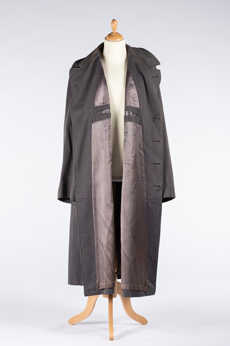 Rett frakk med slag og raglanerme. Foring av silke. Skjult knepping med fire knapper, kan også kneppe slaget under kragen. To lommer med klaff og to innerlommer. Splitt bak med knepping. Spensel nederst på ermene.