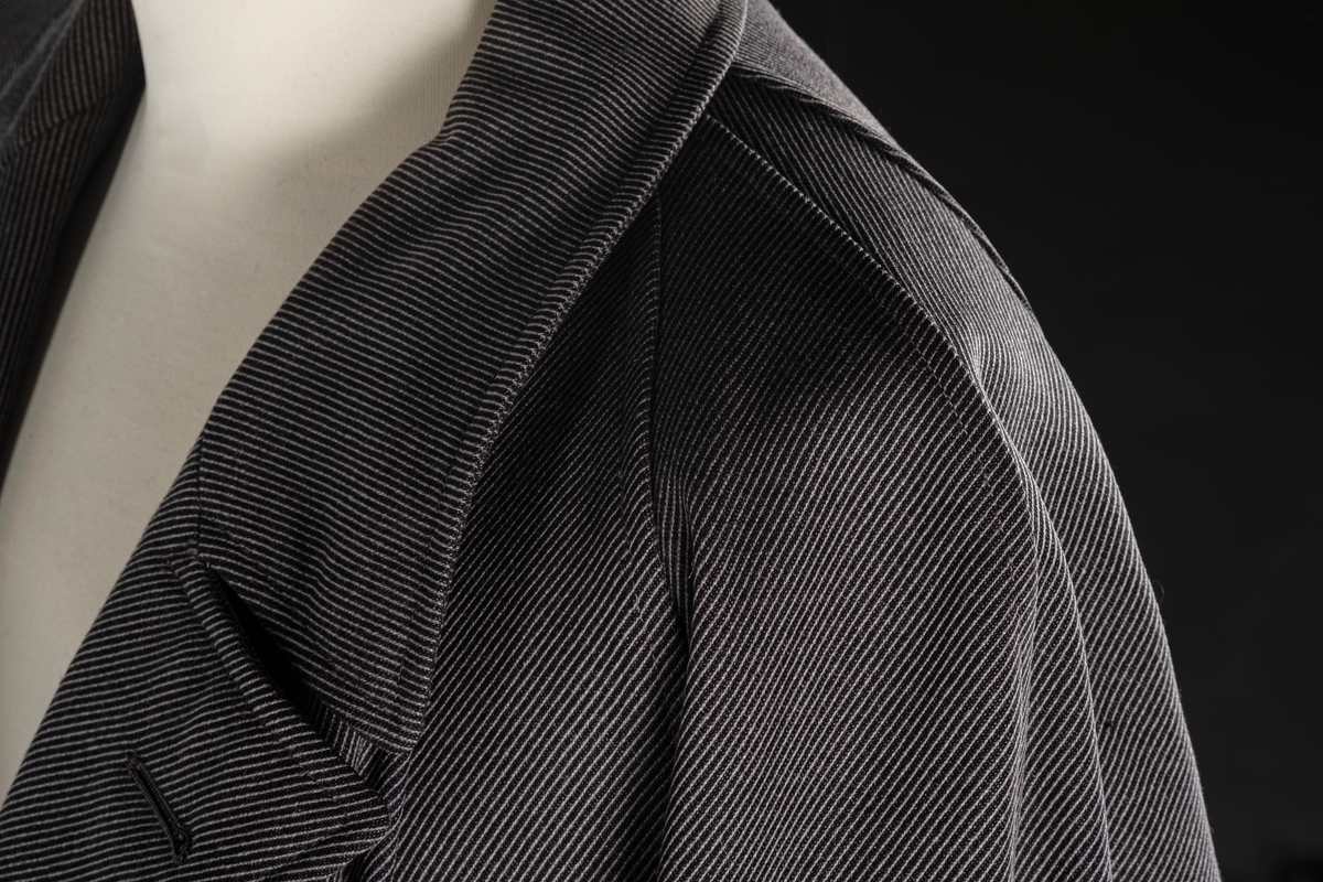 Rett frakk med slag og raglanerme. Foring av silke. Skjult knepping med fire knapper, kan også kneppe slaget under kragen. To lommer med klaff og to innerlommer. Splitt bak med knepping. Spensel nederst på ermene.