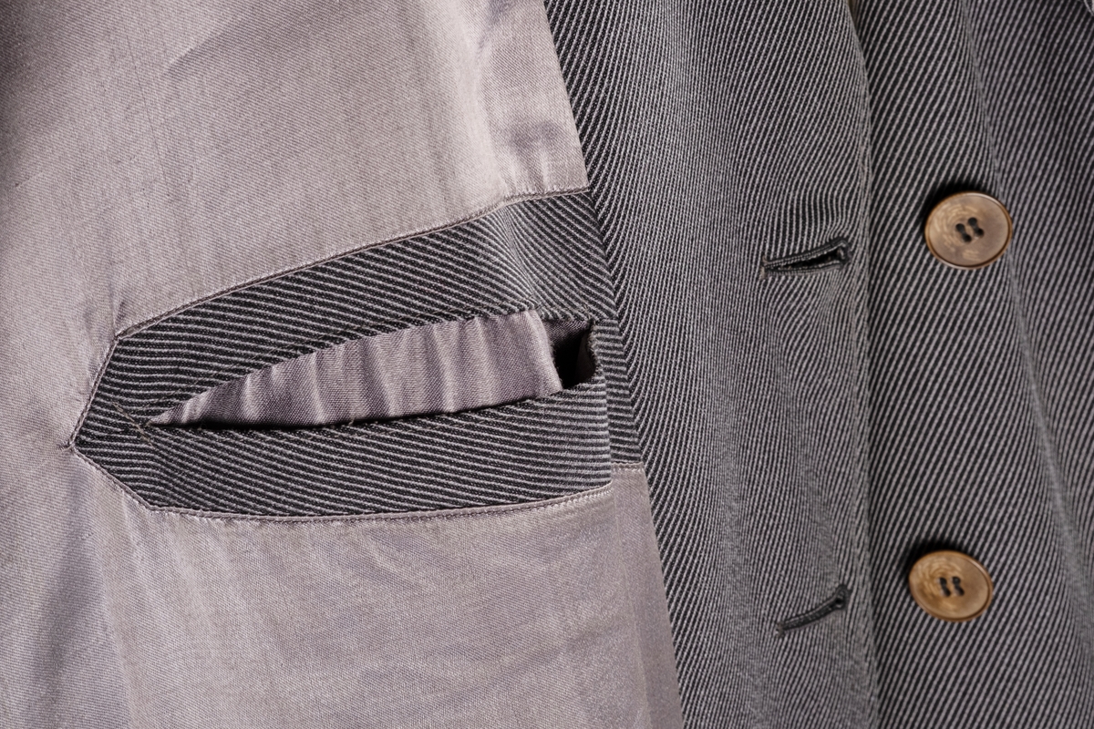 Rett frakk med slag og raglanerme. Foring av silke. Skjult knepping med fire knapper, kan også kneppe slaget under kragen. To lommer med klaff og to innerlommer. Splitt bak med knepping. Spensel nederst på ermene.