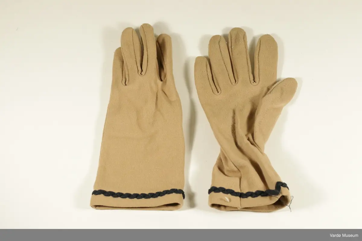 Damehansker til venstre og høyre hånd. Hanskene er beige. De er en sort brodert kant nederst på hansken. Hanskene er tynne. Inni høyre hanske er det sydd inn en lapp hvor det står "Nylon. Made in England". 