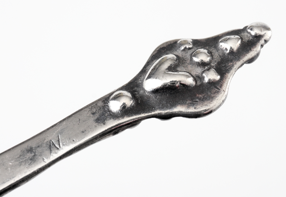 Brännvinssked av silver. 
S.k. kerubsked med kort, platt skaft, dekorerat med hålnät och zick-zackmönster. 
Skaftknopp bestående av ett enkelt (mycket nött) kerubhuvud. Bladet är ovalt med svisselerad blomdekor. 
Stämplar och åldermansranka på skaftets baksida.

Inskrivet i huvudbok 1870