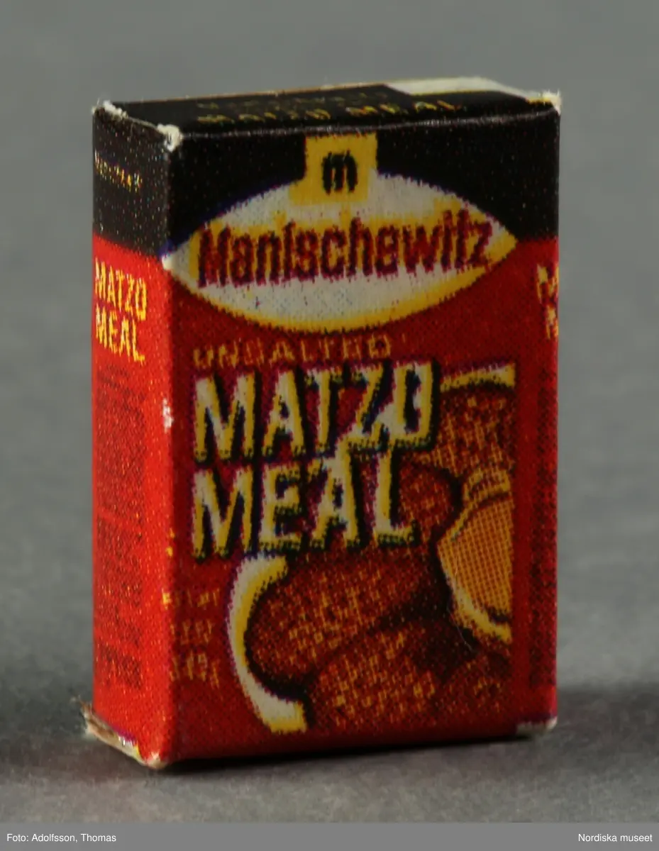 Sju matattrapper föreställande förpackningar med amerikanska och svenska livsmedel som a-b) Corn Flakes (2 st), c) Matzo Meal, d) Quaker Oats, e) Hershey's och f) Marabou mjölkchoklad samt en bit g) hårdost. Hör till inredningen i skafferiet bredvid frukostrummet på första våningen i dockskåp NM.0331721+.