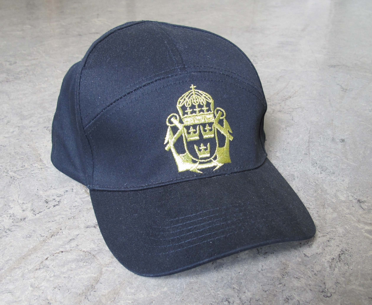 Mörkblå skärmmössa, keps. Framtill emblem i guldfärg: korslagda ankare med tre kronor i krönt sköld.