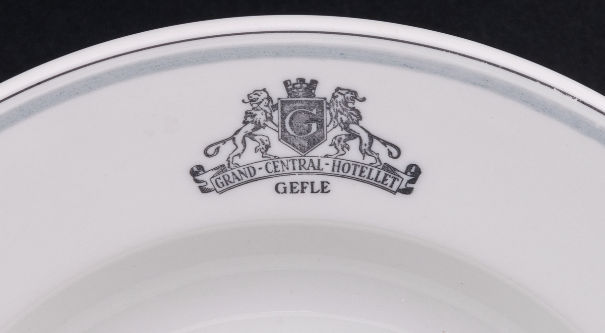 Tallrik, djup. Vit glasyr med ljusgrå och mörkgrå dekor. På brättet emblemet Grand Central Hotellet Gefle, samt två lejon hållande Gävle vapen. Ytterst på brättet två tunna grå ränder.