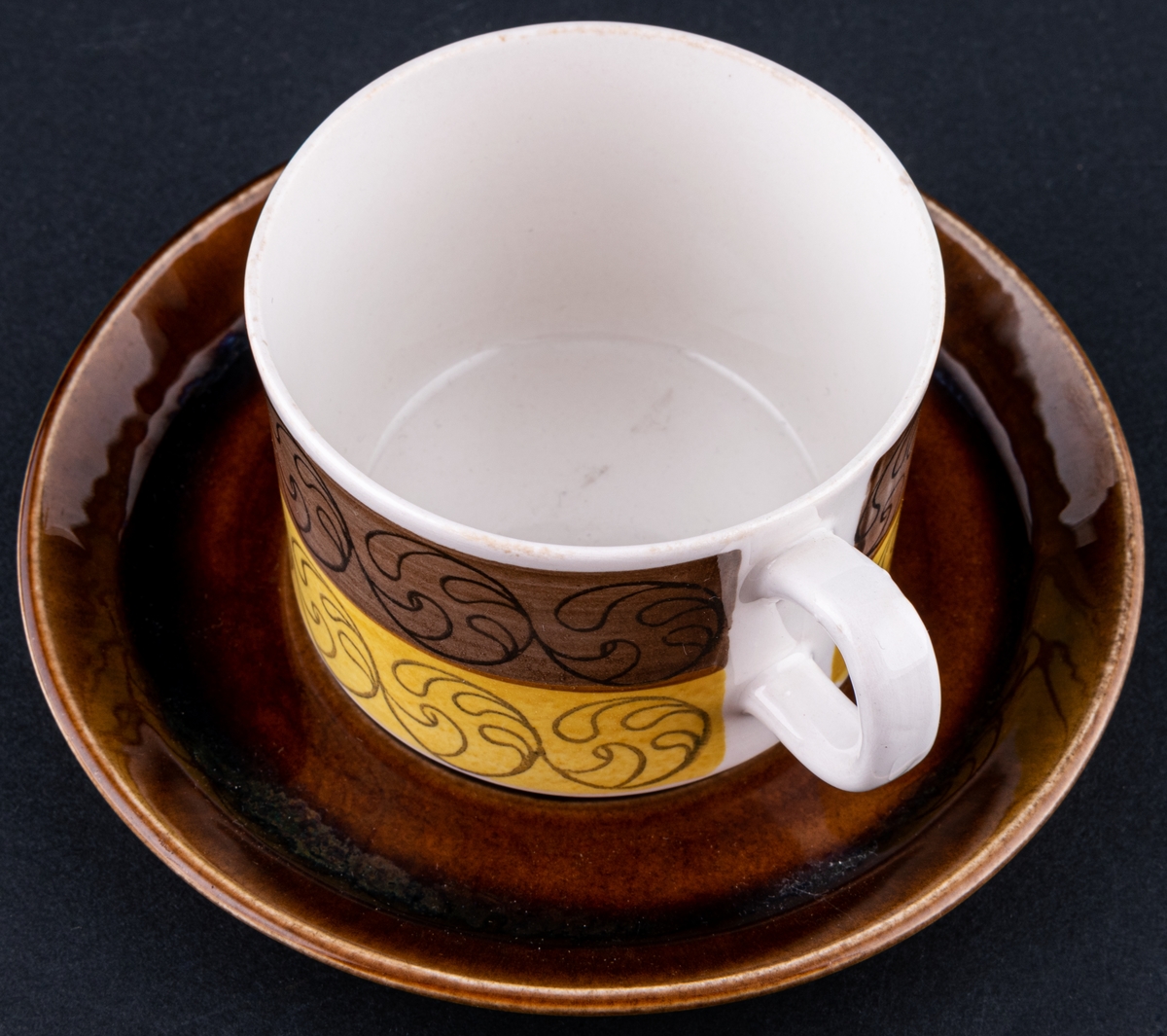 Kaffekopp med fat, modell EH i dekoren Ek. Dekor av Barbro Löfgren-Örtendahl. Runt koppen ett brunt och ett gult band med centrumtryck, fatet helt i brunt.