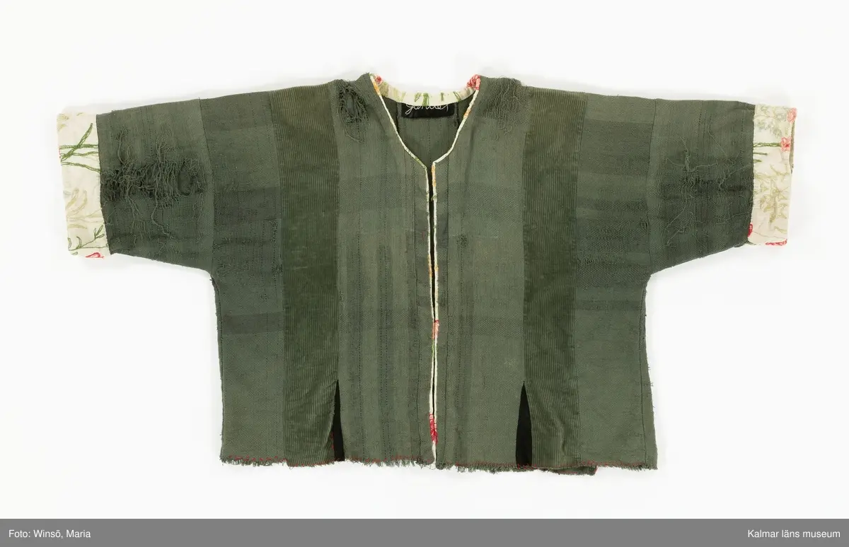 KLM 46157:591. Jacka. För dam. Grön. Rak modell med lång ärm. Sydd av två olika tyger, ett grövre bomullstyg med trådränder i blått och rött samt av bredspårig bomullsmanchester. Jackan är sydd av raka våder. Mellan några av våderna, vid nederkanten, isydda svarta kilar. Öppning mitt fram. Öppning och halsringning kantad med broderat, blommor i röda och gröna nyanser, ljust bommulstyg. Samma tyg är sytt i en bredare avslutning på ärmen. Rå kant vid jackans nederkant, sydd med glesa röda kaststygn. Handbroderad svart etikett på insidan i nacken med texten till stora delar dold av kantremsan. 
Tygerna i plagget är troligen återvunna. Det broderade tyget finns även i klänning KLM 46157:578.