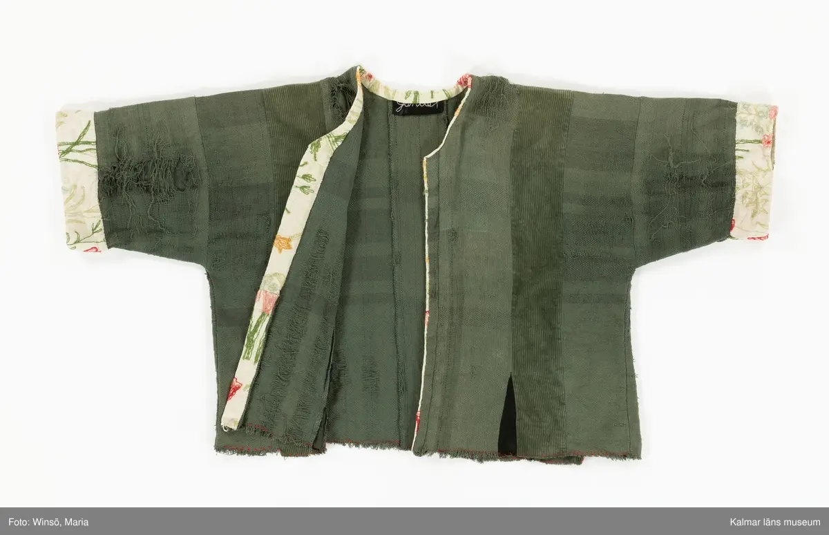 KLM 46157:591. Jacka. För dam. Grön. Rak modell med lång ärm. Sydd av två olika tyger, ett grövre bomullstyg med trådränder i blått och rött samt av bredspårig bomullsmanchester. Jackan är sydd av raka våder. Mellan några av våderna, vid nederkanten, isydda svarta kilar. Öppning mitt fram. Öppning och halsringning kantad med broderat, blommor i röda och gröna nyanser, ljust bommulstyg. Samma tyg är sytt i en bredare avslutning på ärmen. Rå kant vid jackans nederkant, sydd med glesa röda kaststygn. Handbroderad svart etikett på insidan i nacken med texten till stora delar dold av kantremsan. 
Tygerna i plagget är troligen återvunna. Det broderade tyget finns även i klänning KLM 46157:578.