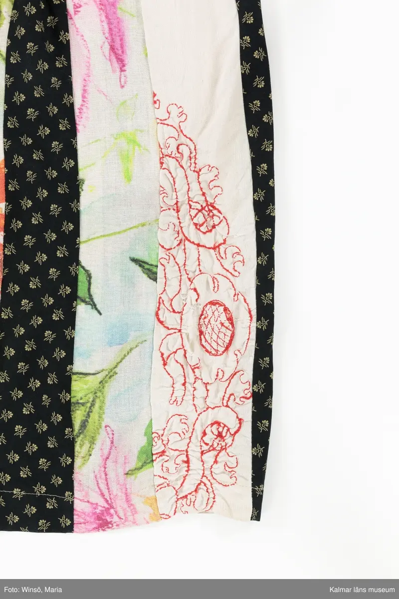 KLM 46157:590. Klänning. Ärmlöst liv av mönstervävt, trianglar och rutor, randigt bomullstyg i rött, rosa, grönt och gult mot svart botten. Halsringning och ärmöppningar kantade på insidan av beige snedslå. Kjol sydd av smala lodräta våder av åtta olika mönstrade tyger, de flesta med tryckt mönster. Tygen är i huvudsak av bomull. Ett tyg har broderat mönster i rött. Ett av de tryckta tygen är formgivet av Lena A Linderholm, mönstret heter Dahlia. Det svarta tyget med små tryckta blommor i gult finns även i kjolen till klänning KLM 46157:588. Det ljusgröna tyget med röda och vita prickar finns i kjolen till klänning KLM 46157:589.