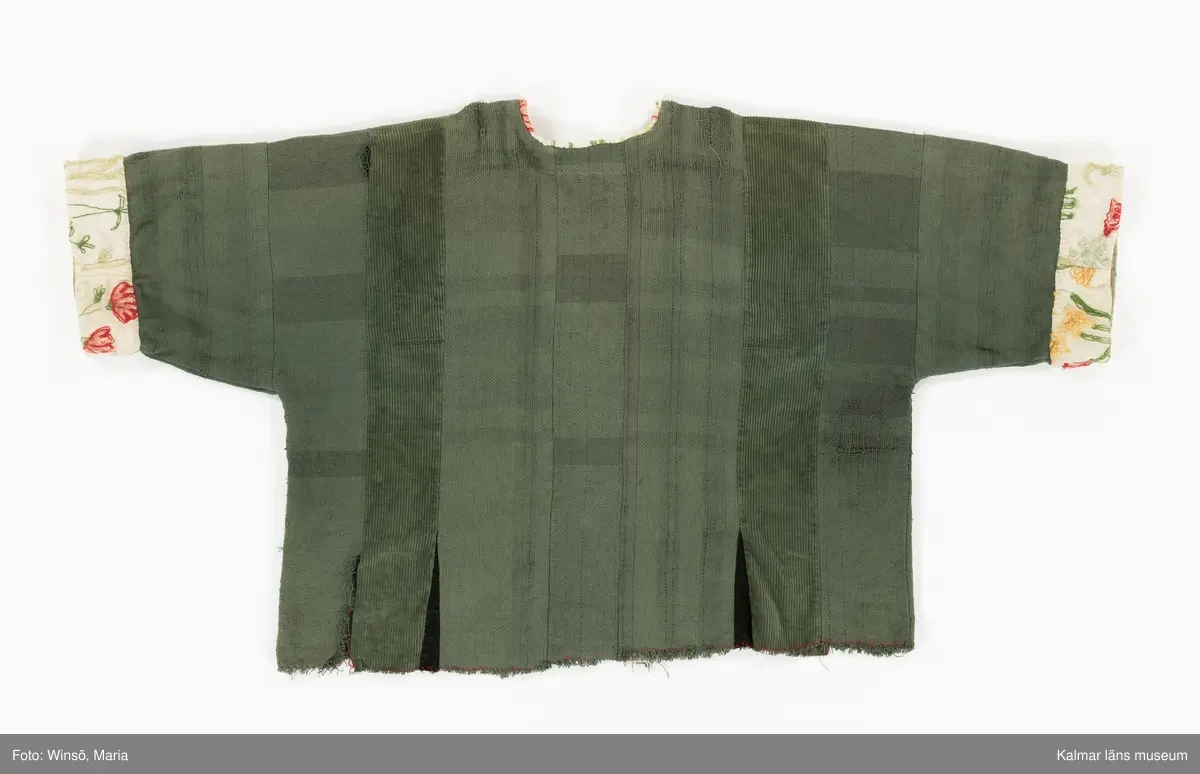 KLM 46157:591. Jacka. För dam. Grön. Rak modell med lång ärm. Sydd av två olika tyger, ett grövre bomullstyg med trådränder i blått och rött samt av bredspårig bomullsmanchester. Jackan är sydd av raka våder. Mellan några av våderna, vid nederkanten, isydda svarta kilar. Öppning mitt fram. Öppning och halsringning kantad med broderat, blommor i röda och gröna nyanser, ljust bommulstyg. Samma tyg är sytt i en bredare avslutning på ärmen. Rå kant vid jackans nederkant, sydd med glesa röda kaststygn. Handbroderad svart etikett på insidan i nacken med texten till stora delar dold av kantremsan. 
Tygerna i plagget är troligen återvunna. Det broderade tyget finns även i klänning KLM 46157:578.