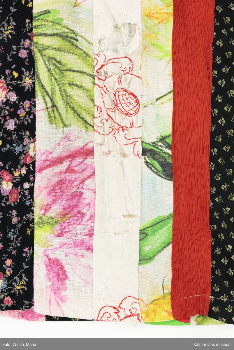 KLM 46157:590. Klänning. Ärmlöst liv av mönstervävt, trianglar och rutor, randigt bomullstyg i rött, rosa, grönt och gult mot svart botten. Halsringning och ärmöppningar kantade på insidan av beige snedslå. Kjol sydd av smala lodräta våder av åtta olika mönstrade tyger, de flesta med tryckt mönster. Tygen är i huvudsak av bomull. Ett tyg har broderat mönster i rött. Ett av de tryckta tygen är formgivet av Lena A Linderholm, mönstret heter Dahlia. Det svarta tyget med små tryckta blommor i gult finns även i kjolen till klänning KLM 46157:588. Det ljusgröna tyget med röda och vita prickar finns i kjolen till klänning KLM 46157:589.