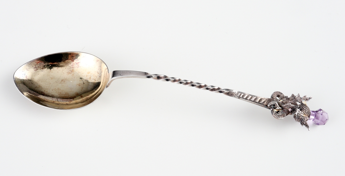Kaffesked i silver.
Skeden har skaftet spiralvridet, avslutat med två ringar, två blad och en tistel. I skaftets yttre ände sitter en
fasettslipad, päronformad ljusviolett glassten. Skedbladet är förgyllt invändigt. 
Stämplar på skaftets baksida.

Inskrivet i huvudbok 1963