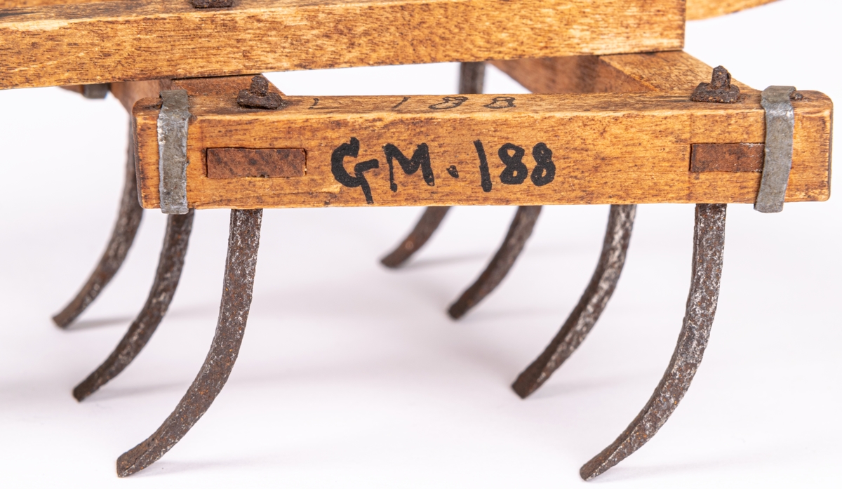 Modell till exstirpator, kallad klösharv, med hjul, åkerbruksredskap. Fyrsidig ram av träbalkar, sture av trä, två framåtriktade åsar, 7 stycken harvjärn i balkramen. Modellens längd 23,5.