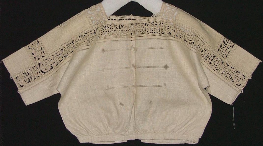 Av grov halvblekt linnelärft, två framstycken, ett bakstycke, besparing, korta ärmar skurna i ett med ovanstående stycken, bårder i spetssöm med grovt halvblekt lingarn kring besparing, hals och på ärmarna, samt hopdragssöm, s.k. myrgång, kring alla kanter samt på ryggstycket, i framkanten åtta sydda tränsar, knappar saknas, blusen rynkad mot smal midjelinning.

Tråckeltråd kvar i midjelinningen, knappar ej isydda.