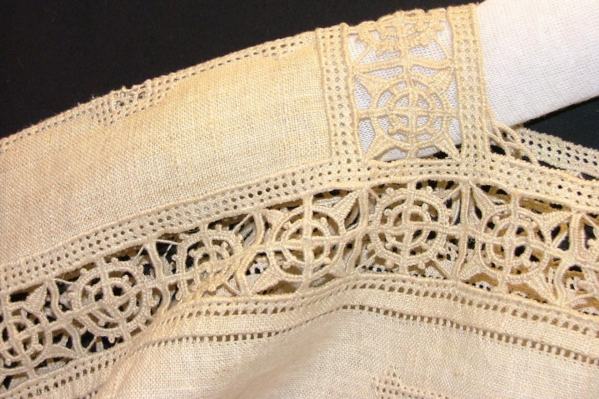 Av grov halvblekt linnelärft, två framstycken, ett bakstycke, besparing, korta ärmar skurna i ett med ovanstående stycken, bårder i spetssöm med grovt halvblekt lingarn kring besparing, hals och på ärmarna, samt hopdragssöm, s.k. myrgång, kring alla kanter samt på ryggstycket, i framkanten åtta sydda tränsar, knappar saknas, blusen rynkad mot smal midjelinning.

Tråckeltråd kvar i midjelinningen, knappar ej isydda.