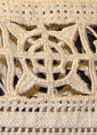 Av grov halvblekt linnelärft, två framstycken, ett bakstycke, besparing, korta ärmar skurna i ett med ovanstående stycken, bårder i spetssöm med grovt halvblekt lingarn kring besparing, hals och på ärmarna, samt hopdragssöm, s.k. myrgång, kring alla kanter samt på ryggstycket, i framkanten åtta sydda tränsar, knappar saknas, blusen rynkad mot smal midjelinning.

Tråckeltråd kvar i midjelinningen, knappar ej isydda.