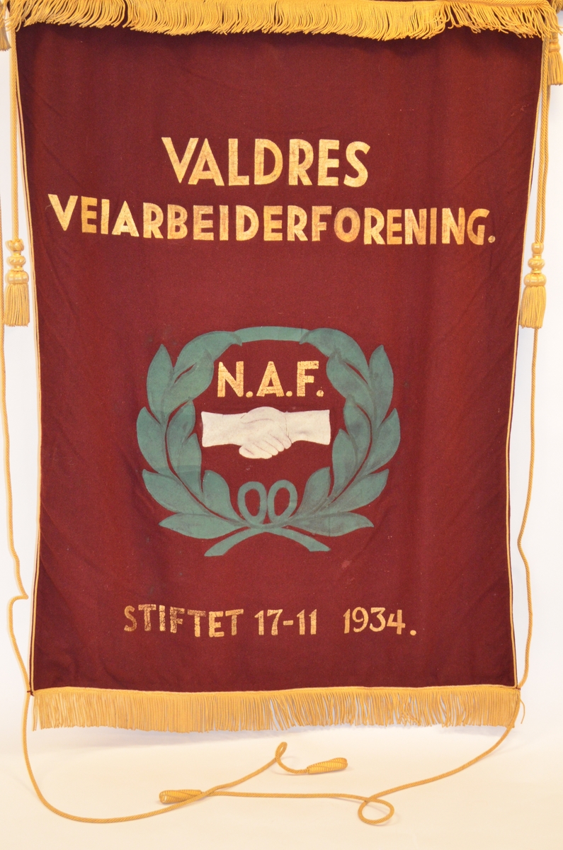 Fane i burgunder raudt ulltøy, lagt dobbelt. Teksten på fana er måla på med gullfarge. Dei andre motiva (bladkransen og hendene) er også måla på tøyet. Kanten i øvre- og nedrekant er påsett gullfarga frynse-bord. Langs sidene er det gullsnor med duskar. På baksida er teksten også måla på med gullfarge. 
Opphengt i øvre i kanten med ei gullmåla trestang med dreia kuler i kvar ende. Denne er tredd inn i ein løpgang. Krok i metall midt på stanga.