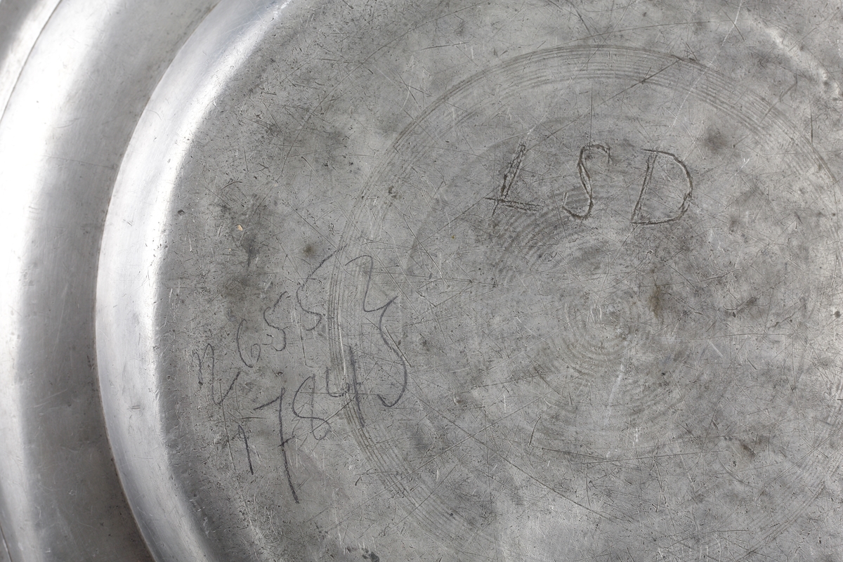 Fat av tenn. Rund, djup modell med uppsvängd bräm, dekorerad med en svarvad linje. 
Ägarinitialer på bottnens utsida: "LSD". Stämplar (mycket nötta och svårtydda) i fatets spegel.

Inskrivet i huvudbok 1903.