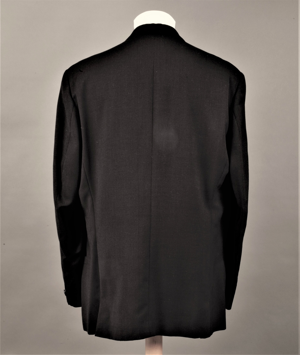 Hoftelang, dobbeltspent, med 6 metallknapper, med Versace-husets "Istante"-logo - Zevs hode - i relieff. Lange slag, og kun de nederste 4 knapper skal kneppes. Lapellene (slagene) er spisse og lengre enn kragen. Ett ekstra knapphull for innvendig knapping, men knappen på innsiden av venstre forstykke mangler. To-søms ermer med falsk splitt, lukket knapphull og pynteknapp. Horisontal lomme med belegg på hver hofte og stikklomme med belegg på venstre bryst. Innvendig, passepoilert lomme på hvert bryst, den høyre lukket med klaff og usynlig knapp. Søm midt bak og splitt nederst i hver panelsøm bak. Forstykkene er skåret rettvinklet i nederkant midt foran. Helfôret med svart fôrsilke. Kragestanden og kragen er kledd med filtaktig materiale på baksiden og jakken har puter i skuldrene.